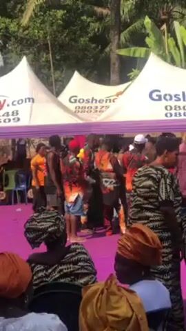 The video shows an outdoor event, possibly a celebration, under white tents with purple trim and the name "Goshencity" printed on them. People are dressed in colorful, patterned clothing, gathering and moving around. Some are dancing, while others are interacting near tables and chairs. The scene is filled with activity and the sound of music or talking is implied.  The camera moves, showing different parts of the event and the attendees. The environment is sunny, with trees visible in the background.