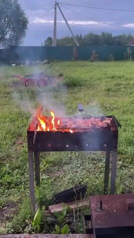 The video shows an outdoor scene at dusk, focusing on a charcoal grill with flames and smoking meat. The grill is positioned in a grassy yard, with trees and a fence in the background. A power line pole is visible in the distance. In the middle ground, a red wheelbarrow is overturned in the grass. The lighting suggests it is early evening or late afternoon.