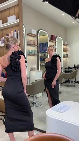 A woman with blonde hair pulled back in an updo, wearing a black one-shoulder dress with ruffled detailing on the shoulder and a thigh-high slit, poses in front of a mirror in what appears to be a salon. She is reflected in the mirror, showing both the front and back of the dress. The salon environment includes shelving units stocked with hair products, salon chairs, and mirrors with illuminated borders. A white table with a document on it is visible in the foreground, and a light fixture is positioned above the scene. The woman is looking at her reflection and adjusting her pose.