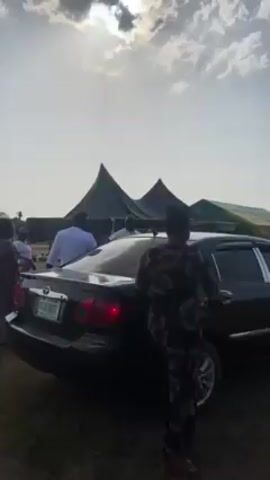A woman in a long black floral dress enters the back seat of a black Toyota sedan parked in a grassy field. The car has a government license plate. In the background, there are several black tents and a few small buildings. The sky is overcast with a bright sun breaking through the clouds. Other people are visible near the tents.