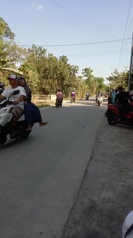 The video displays a street scene from a low-angle, landscape perspective, oriented vertically. A paved road runs diagonally across the frame, bordered by a rougher, grey surface on the right and a grassy area with trees and vegetation on the left. Several motorcycles are present, some parked and others in motion. On the left side of the road, multiple people are gathered, some on or near motorcycles. One person is wearing a blue shirt with text on the back. On the right side of the road, a different person is partially visible, wearing a blue shirt and jeans, facing away from the camera. The sky is clear and blue. The overall impression is of a gathering or a moment of activity on a sunny day.