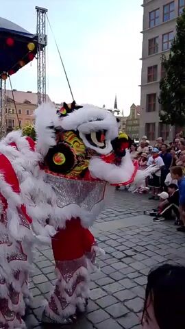 A group of people are gathered in an outdoor square paved with cobblestones. A stage with a large screen and metal scaffolding is visible in the background. Two lion dancers, wearing elaborate red and white costumes with shimmering accents, are performing. One lion dancer is positioned in the foreground, facing left, with its head held high. The other lion dancer is slightly further back and to the right, also facing left. A crowd of spectators, including adults and children, are watching the performance, some sitting on the ground. The architecture of surrounding buildings suggests an urban setting. The sky is clear and the lighting indicates daytime.