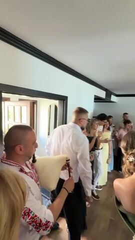 A man in traditional clothing plays bagpipes at a wedding. Guests, dressed in formal attire, stand in a hallway or room, some clapping. A man in a white shirt and black pants walks away from the camera. The room has white walls with black trim and wooden floors. There are doorways leading to other areas and a decorative mural on the far wall. The atmosphere suggests a celebratory event.