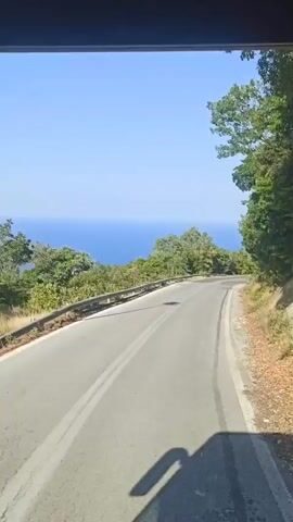 This media file shows a view from inside a vehicle traveling on a winding road. The road curves to the right, with a guardrail on the left side. Beyond the guardrail, lush green trees and vegetation cover a hillside that slopes down towards a body of water. The water, appearing to be the sea, is a deep blue and stretches to the horizon under a clear, bright blue sky. The sun is high, indicating it is likely daytime, and the weather appears to be clear and sunny. The scene suggests a scenic drive, possibly in a mountainous coastal region.

Notable details include the texture of the asphalt road, which shows some wear and tear with patches and cracks. Shadows from the vehicle are cast on the road surface, and tire marks are visible on the right edge of the pavement. The foliage is dense and vibrant green, with some trees reaching higher than others. The overall impression is one of a beautiful natural landscape on a warm, sunny day. There is no visible text in the media file. The context provided states the location is Palaios Panteleimonas, Greece, which aligns with the visual cues of a coastal, mountainous region with dense vegetation.