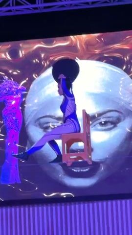 A performer in a blue and white bodysuit is sitting on an orange chair, balanced on a tightrope. The performer is facing left, with their right arm extended upwards. Behind the performer, a large projection displays a face with eyes looking towards the viewer, and swirling orange and purple patterns. To the left of the projection, a purple, illuminated figure stands upright. A man in a black t-shirt and black pants is standing on the right side of the frame, watching the performance. The stage is lit with purple lights and the background features a striped pattern.