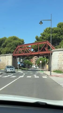 The video captures a car driving forward on an asphalt road towards a red metal bridge. The car stops briefly before passing under the bridge. The road has a painted white arrow pointing straight and a stop line. A yield sign is visible on the right, and a "turn left" sign is on the left. Green trees line both sides of the road, and residential buildings are visible in the background. The sky is clear and blue.