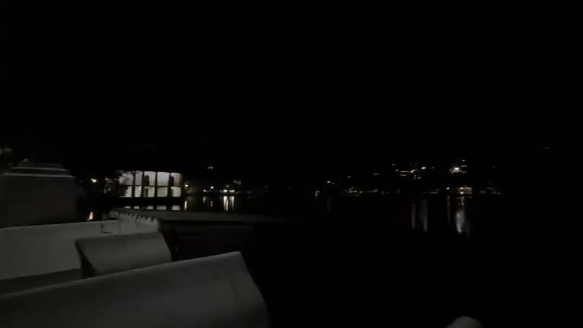 The video shows a dark night scene with a body of water in the foreground and a town or city visible in the distance. The lights from the town are reflected in the water. In the foreground, there are some white, low-backed chairs. The overall impression is one of darkness and tranquility. The camera is relatively still.