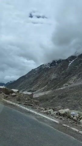 A video taken from a vehicle traveling along a mountain road. The road is paved and curves to the left. To the right of the road, there is a steep, rocky mountain slope. Several concrete barriers are visible along the side of the road, separating it from the slope. The mountain is covered in loose scree and some patches of snow are visible near the summit. The sky is overcast with heavy clouds, obscuring much of the mountain's upper reaches. The overall impression is one of a remote, rugged mountain environment.