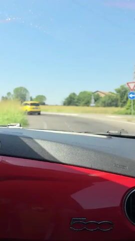 The video begins with a view from inside a red car, looking out the windshield at a rural road on a sunny day. The dashboard of the car is visible in the foreground, featuring the "500" logo. In the distance, a yellow truck is driving down the road. The car is moving, as indicated by the slight motion blur. The road is flanked by green fields and trees, and a clear blue sky is above. The yellow truck continues to drive away from the camera, eventually disappearing from view as the car continues its journey. The video ends with a similar view of the road and the environment.
