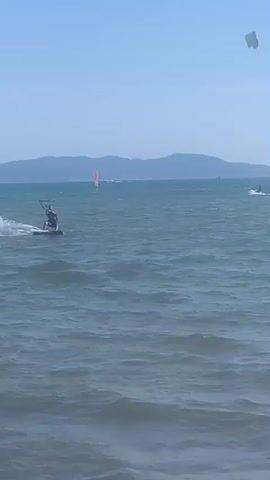 The video shows a bright, sunny day with a clear blue sky over a body of water. In the foreground, the choppy surface of the water is visible, with small waves breaking. In the middle ground, a person is kitesurfing. They are on a board, being pulled by a kite in the air. The kitesurfer is leaning back, controlling the kite and the board. White spray is visible behind the board, indicating movement. There are other kitesurfers and sailboats in the distance, scattered across the water. The horizon is marked by a distant, hazy landmass with a few structures visible on it, possibly buildings or a castle. The overall scene depicts a recreational activity taking place in an open-water environment under favorable weather conditions.