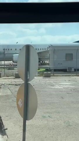 The video shows an airplane with the text "IAI ELTA" on its side. The airplane appears to be parked at an airport or airfield. There are various structures and equipment around the airplane, including scaffolding, stairs, and barrels. A red car is visible parked near the airplane. The scene is viewed through a fence, suggesting the video was taken from a restricted area. The sky is cloudy, and the overall lighting suggests it is daytime.