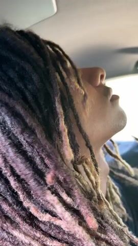 A person with multi-colored dreadlocks (brown, blond, and reddish/pinkish tones) is filmed from a low angle inside a vehicle. The person's head is tilted back, with their eyes seemingly closed or looking upwards, and their mouth slightly open. Their long dreadlocks are flowing and moving, suggesting the vehicle is in motion, possibly with an open window or sunroof. The background is blurred, indicating movement. The distinct sound of wind rushing past is audible, along with faint background music. The lighting suggests it is daytime. The person appears relaxed. There is no visible text.