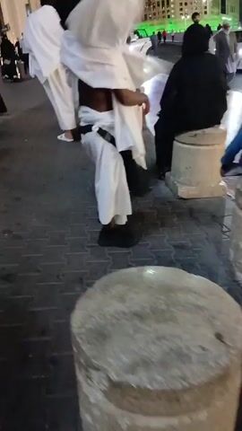 The video shows a street scene at night in what appears to be a Middle Eastern city. People are walking along a sidewalk, some dressed in traditional Islamic clothing, including white robes and black abayas. There are tall buildings in the background. A few people are standing near the curb. The camera is handheld and moves with the person filming, showing the surrounding area. Some streetlights are visible.
