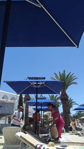 A group of people are gathered near a swimming pool. There are several blue umbrellas providing shade, and palm trees are visible in the background. Some people are sitting on lounge chairs, while others are standing. One woman in a pink dress is interacting with a straw bag. Another man is wearing a white t-shirt and appears to be looking at something on the ground. The sky is clear and blue.