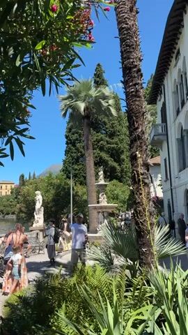 The media file captures a sunny day scene in Varenna, Italy, with a focus on a picturesque waterfront garden. Several people are present, appearing to be tourists enjoying the scenic view and the garden's ambiance.

In the foreground, lush green foliage and a large palm tree dominate the frame, with sunlight filtering through the leaves. A pathway is visible, with a few individuals standing and interacting. Among them, a group of women and a child are gathered near a stone railing overlooking a body of water, likely Lake Como. One woman is wearing a colorful floral dress, another a white dress, and a child in shorts and a t-shirt. A man in a white t-shirt and shorts is also visible, possibly a guide or a member of staff.

The background reveals a stunning lakeside landscape. The clear blue water of Lake Como stretches out, with the distant shore dotted with buildings and verdant hills. A yellow villa stands out on the left side of the frame. Several classical-style statues and a fountain are placed throughout the garden, adding to the aesthetic appeal. The overall atmosphere is relaxed and pleasant, suggesting a leisurely vacation or sightseeing activity.

The time of day appears to be midday, indicated by the bright sunlight and clear blue sky, with no clouds visible. The weather seems warm and inviting. The presence of palm trees and the style of the architecture suggest a Mediterranean climate.

There is no visible text in the media file. The scene evokes a sense of tranquility and appreciation for natural beauty and historical surroundings.