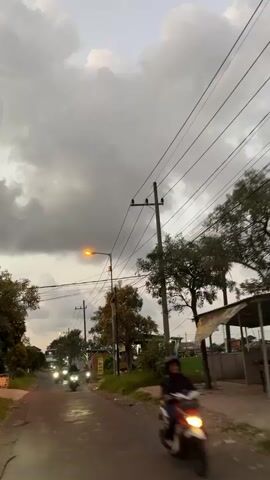 The video captures a low-angle, first-person view from a vehicle traveling on a paved road in Wunut, Indonesia. The time of day appears to be late afternoon or early evening, as indicated by the dim, diffused lighting and the streetlights beginning to illuminate. The sky is heavily overcast with thick, grey clouds, suggesting the possibility of rain or a storm.

The main subjects are two motorcycles traveling ahead on the road, their headlights brightly lit against the darkening sky. The road itself is bordered by vegetation, including trees and grass. Electrical poles with numerous wires stretch across the frame, a common sight in many urban and semi-urban environments. In the background, glimpses of simple buildings and a green field can be seen. On the right side of the road, a small structure with a slanted roof, possibly a roadside stall or shelter, is visible.

The overall atmosphere is somewhat muted due to the weather conditions, but the bright headlights of the motorcycles and the single illuminated streetlight offer points of visual interest. There is no visible text in the media file. The scene evokes a sense of everyday life on a road in Indonesia, with the weather creating a slightly dramatic backdrop.