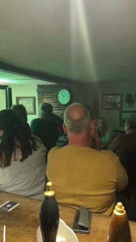 A dimly lit bar with blue, purple, green and red stage lighting shows a group of mostly men dancing in front of a band or entertainment.  The camera focuses on the backs of people seated at the bar, including a bald man in a yellow sweater. Several beer bottles are on the bar. A window with white curtains is in the background. Artwork hangs on the walls.