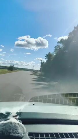The video shows a view from the inside of a car looking out the windshield. The car is driving on a paved road through a rural landscape under a bright blue sky with scattered white clouds. To the right of the road, there are trees and vegetation, and to the left, there is a large green field. In the distance, there are more clouds and what appears to be a cluster of buildings. The sun is visible in the upper right corner, casting a glare on the windshield. A small, fuzzy grey object is attached to the dashboard in the lower left corner. The camera appears to be mounted on the dashboard.