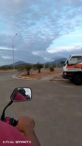 The video shows a first-person perspective of someone riding a motorcycle on a paved road. The sky is blue with scattered clouds, and there are mountains visible in the distance. The road has palm trees lining one side, and there is a sidewalk and curb separating the road from the palm trees. There is a white truck parked on the side of the road in the opening shot. The camera is mounted on the motorcycle and the ride appears smooth. The side mirror of the motorcycle is visible in the lower left corner of the frame. A brief glimpse of another motorcycle passing on the other side of the road is seen around halfway through.  The video is labeled "Infinix SMART 8" at the bottom of the frame.