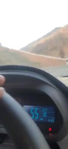 The video shows a view from inside a car, looking out the windshield. The car is on a road that appears to be in a mountainous or hilly area. The road has a guardrail on the right side. In the distance, there are hills with dry, sparse vegetation. The sky is clear and bright.

The dashboard of the car is visible in the foreground. A steering wheel is partially visible on the left. A digital display on the dashboard shows the number "54", likely indicating the car's speed in kilometers per hour, and "22C", indicating the outside temperature in Celsius.

A white truck with a trailer passes by in the distance on the road. The trailer is yellow with black lettering that is not clearly legible. The truck is traveling in the opposite direction to the car. The video appears to be taken while the car is in motion.