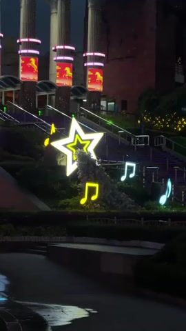 The video showcases an outdoor illuminated display at night, featuring a tiered landscape with musical note and star-shaped light figures. In the background are structures with illuminated columns and images. Statues are positioned atop one of the upper tiers. The display includes neon signage, including Chinese characters and the English words "The World." A water fountain is also active in the foreground. The environment appears to be urban, with buildings and cranes visible in the distance. The overall scene suggests a festive or celebratory atmosphere.