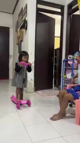 A young girl wearing a gray dress and black long-sleeved shirt stands on a pink scooter in what appears to be a home interior. She is looking at another girl sitting on a small pink chair. The room has white tile floors, and the background shows a door, a bookshelf filled with items, and framed pictures on the wall. The girl on the scooter adjusts the handlebars and appears to be preparing to ride. The other girl watches her. An adult man is seen briefly in the background.