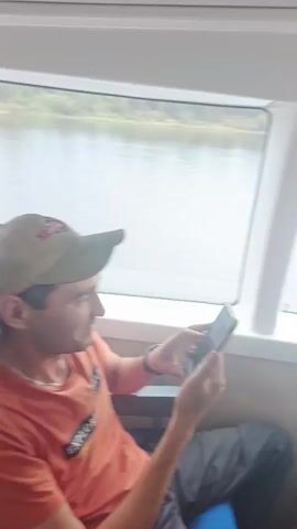 The video shows the interior of a passenger boat or ferry. Several passengers are visible, some looking out the windows at the water and scenery, while others are looking at their phones or talking. One person is holding a phone and appears to be recording or taking pictures. Another person is looking at a tablet. Two women are standing and looking out a window together. Another person, a man, is standing and looking out a window. A television screen is mounted on the wall, showing an image. The seating appears to be arranged in rows, with blue and light-colored seats. The windows offer a view of a body of water and a cloudy sky. The overall atmosphere seems relaxed, with people engaged in their own activities during the journey.