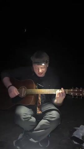 A young man sits cross-legged in the dark, playing an acoustic guitar. He wears a headlamp that illuminates his face. He is dressed in a dark jacket, grey pants, and black and white sneakers. He has a guitar strap with a colorful pattern. The background is completely dark.