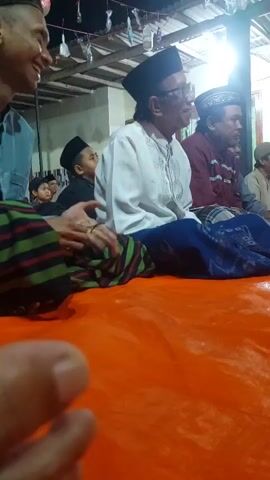 The video shows a group of men sitting on an orange tarpaulin. One man in the foreground is wearing a light blue shirt and a patterned sarong. He is smiling and looking to the left. In the center of the frame, a man wearing a white shirt and a black peci is seated, looking to the right. Behind him, other men are also seated. The environment appears to be an outdoor or semi-outdoor gathering, possibly at night, with a roof structure visible in the background and some decorations hanging above. The lighting is dim but sufficient to illuminate the faces of the people. The camera perspective is low, as if held by someone seated on the tarpaulin.