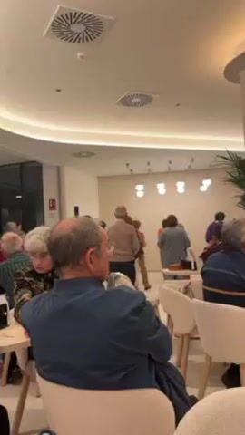 The video shows a brightly lit indoor space, likely a hotel lobby or common area, with a mix of people engaging in different activities. In the foreground, several people are seated in cushioned chairs around small tables, some watching a group of others dancing near a wall decorated with hanging lights and greenery. The dancers appear to be older adults. In the background, another group of people is gathered around a television screen watching a sporting event. The camera pans slightly, showing more of the interior space and the people within it. Overall, the atmosphere seems relaxed and social, with people enjoying different forms of entertainment and conversation.
