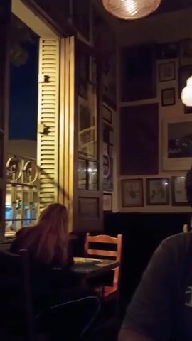 The scene appears to be inside a restaurant with dim lighting. Several framed pictures and posters adorn the yellow walls, hung in a collage style. A ceiling fan is visible.  Two people are seated at tables. One person with dark hair is closest to the camera, wearing a greenish-grey shirt, and is looking down, perhaps at a table. In the background, another person is seated at a table further away. A doorway leads into another section of the restaurant. Light fixtures are hanging from the ceiling, providing soft illumination. A wooden door with window panes is visible on the right side of the shot.