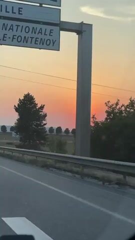 The video shows a sunset view from a vehicle. The sun, a bright orb, is partially obscured by a tree in the foreground and is descending towards the horizon. The sky is a gradient of orange and pink hues. A road with a metal guardrail runs diagonally across the lower part of the frame. In the background, there is a field with a line of trees and some signs, including a speed limit sign indicating 70. Another sign appears to be a roundabout symbol. The overall scene depicts a tranquil rural landscape at dusk.