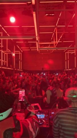 The video shows a crowded nightclub. The perspective is from the DJ booth looking out at the dance floor. Red light floods the entire space. Numerous people in the audience are holding up cell phones, presumably recording the performance. A DJ is working at a Pioneer mixer. Two other people are also visible behind the DJ station. Overhead, the ceiling structure is visible as a grid of lighted white tubes.