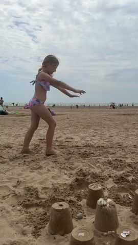 A young girl in a colorful swimsuit is performing a gymnastic move on a sandy beach. She is captured in mid-air, with one leg extended upwards and the other bent, while her hands are on the sand for support. The scene is set on a beach with a cloudy sky overhead. In the background, other beachgoers can be seen enjoying the day, with umbrellas and tents dotting the shore. There are also sandcastles in the foreground, suggesting children's play. The overall atmosphere appears to be one of a leisurely day at the beach. The girl's posture and the movement suggest she is engaged in an athletic or playful activity. The presence of the sea and other people in the distance indicates a public beach location. There is no visible text in the media file.