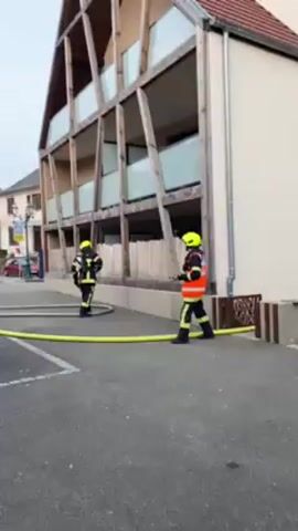 The video shows two firefighters in full gear, including yellow helmets, walking next to a yellow fire hose on a gray paved road in what appears to be a small town or village. They are heading toward a modern, multi-story building with a unique wooden frame design around its balconies. A third firefighter emerges from the open door of a red and yellow fire truck, also in full gear. The scene is set on an overcast day. There is a street sign and other buildings visible in the background.