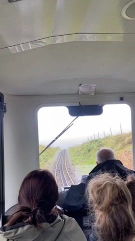The video shows the view from inside a small train car traveling up a hill on a narrow gauge railway. A man in the driver's seat is operating the train, while two female passengers sit behind him. The weather is overcast and rainy, with a wiper blade clearing the window. The tracks lead up a grassy hillside towards the horizon. A warning sign is visible on the roof of the car.