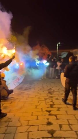 At night, a group of young people are gathered on a brick walkway, holding and waving smoke bombs and sparklers. The smoke is white, orange, and blue. They are positioned around a banner, partially visible, that has writing on it. The sky is dark, and a street light is in the distance. Some people are wearing dark jackets. One person is hitting a tambourine. The group appears to be celebrating or protesting.