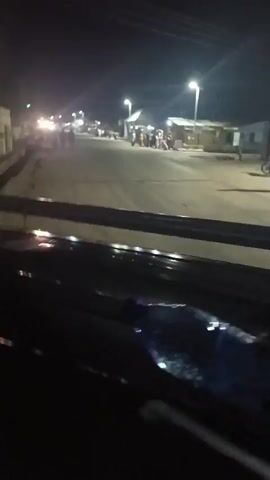 The video shows a night scene from inside a vehicle driving down a street. The street is lit by streetlights and the headlights of other vehicles. There are people walking on the sidewalks and a motorcycle driving on the street. The buildings along the street appear to be simple structures. There is a drainage ditch running alongside the road. The overall impression is a typical small town or village scene at night.