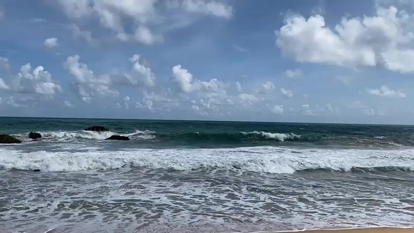 A medium shot shows a view of the ocean from a beach. The waves are breaking, creating white foam. The sky is blue with scattered white clouds. The water is a deep green and blue color. The sand is visible in the foreground. The scene is calm and serene.