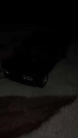 A single, black, die-cast model car is centrally framed on a white surface with brown patterns. The lighting slowly brightens to fully illuminate the car, revealing its Lada-like design and silver-colored rims. The scene is still and focused solely on the miniature vehicle.