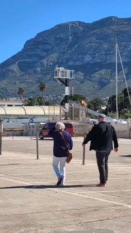 Two people, a man and a woman, are walking away from the camera on a paved area marked with parking lines. The woman is wearing a blue sweater, white pants, and carrying a brown bag. The man is wearing a dark jacket, dark pants, and a cap. In the background, there is a large mountain, a red van, a structure with lights, and a building with a curved roof. The sky is clear and blue. A few palm trees are also visible. The scene appears to be at a port or marina.