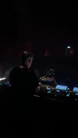 The video shows a DJ performing at a nightclub. The DJ is in the foreground, facing away from the camera, with a setup of turntables and controllers. A crowd of people is visible in the background, dancing and enjoying the music. The atmosphere is dimly lit with colored lights and fog, creating a typical nightclub scene. The camera pans slightly, showing different angles of the DJ and the crowd.