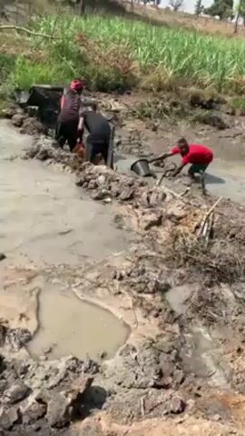 The video shows three people working in a muddy, waterlogged area. One person in a red shirt and blue shorts is using a bucket to scoop water out of the mud. Two other people, one wearing a red head covering and carrying a yellow container, are also working in the mud. The environment appears to be rural, with green vegetation in the background.