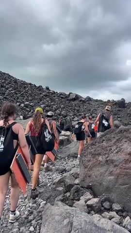 In this media file, a group of people are hiking up a rocky, barren slope. The individuals, dressed in casual hiking attire, carry backpacks and some are carrying large, flat objects, possibly boards or panels. The terrain is dominated by volcanic rocks and scree, suggesting a volcanic landscape. The sky above is overcast with dramatic, dark clouds, indicating a potentially moody or stormy weather condition. The overall atmosphere is one of exertion and adventure as the group navigates the challenging terrain. The specific location is Rota, Nicaragua, known for its volcanic geography. The time of day appears to be daytime, given the natural light, but the cloudy sky might obscure direct sunlight. The image captures a moment of upward movement and shared effort amongst the hikers.