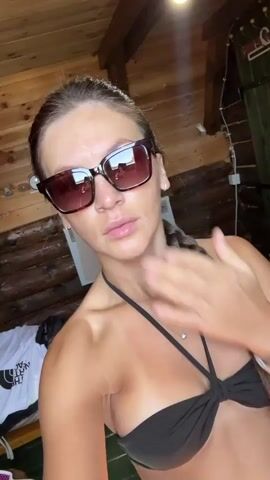 A woman with long brown hair braided to her right wears sunglasses and a black bikini top. She is indoors in a room with log walls and wooden ceiling beams. She holds her right hand to her mouth and then blows a kiss towards the camera.