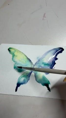 The video shows a close-up view of a watercolor painting of a butterfly. The butterfly has its wings spread open and is depicted in shades of blue, green, and yellow. A paintbrush with a gray handle and white bristles is being used to blend the colors on the left wing of the butterfly. The brush is held by a person whose hand is visible on the right side of the frame, with a bright yellow fingernail. The painting is on a white piece of paper, which is placed on a white surface. There are faint pink splotches of paint visible on the white surface above the butterfly. The video captures a single, continuous action of painting.