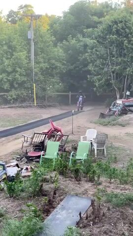 A person on a dirt bike approaches a ramp. The dirt bike launches off the ramp and into the air. The person performs a trick on the dirt bike while in the air. The dirt bike lands on an inflatable landing pad. The video is filmed outdoors during the day with trees and a clear sky in the background.