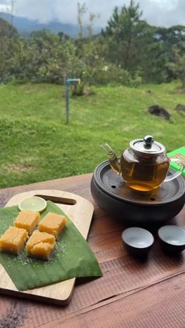 A medium shot shows a glass teapot with a silver lid filled with amber-colored liquid on a black base, situated on a wooden table. Beside the teapot are two small, dark bowls. To the left of the teapot, on a wooden cutting board covered with a green leaf, are four square, light-yellow sweets dusted with white powder and a thin slice of lemon. In the background, a lush green landscape unfolds under a cloudy sky, featuring rolling hills, trees, and a distant view of mountains. A blue pipe and a small structure are visible in the mid-ground. The video zooms out slightly, panning away from the table and the food, to focus more on the expansive outdoor scenery.