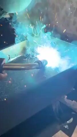 A person is welding two pieces of metal together in a bright, sparking display. The welding torch is held in gloved hands, producing a vivid blue and white light with orange sparks flying around the welding point. The metal is a dull, dark color. The background is dark and indistinct, with the focus clearly on the welding process. The camera is positioned relatively close to the action, providing a detailed view of the welding activity.