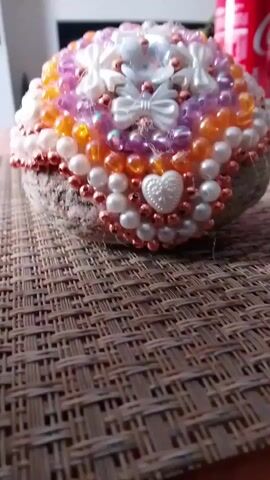 A close-up shot focuses on a rounded object covered in decorative beads and small bows. The beads are various sizes and colors including white, pink, orange, and iridescent purple, with some metallic rose gold beads mixed in. There are several small white bows and a few white heart-shaped embellishments. The object appears to be resting on a textured surface that looks like woven matting. In the background, a red and white can is visible, and the lighting suggests an indoor setting. The camera is steadily focused on the beaded object, slowly panning across its surface.